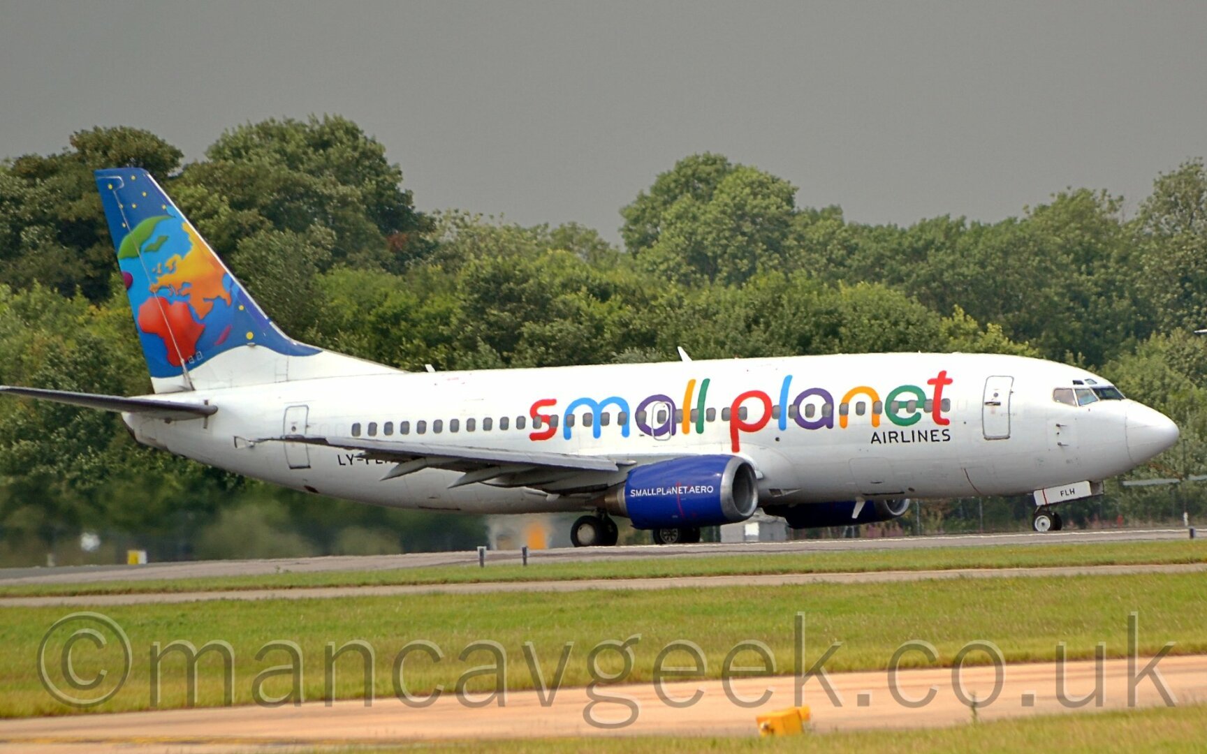 White jet airliner with colourful "Small Planet" titles and a cartoon image of a globe on the tail at an airport, with trees in the background.