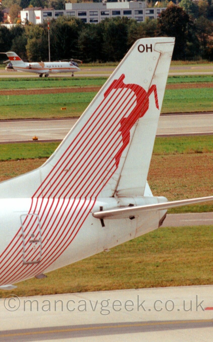 Close up of the tail of a white jet airliner with a stylised image of a  leaping gazelle or similar animal. In te background is a bizjet with a Swiss flag on the tail