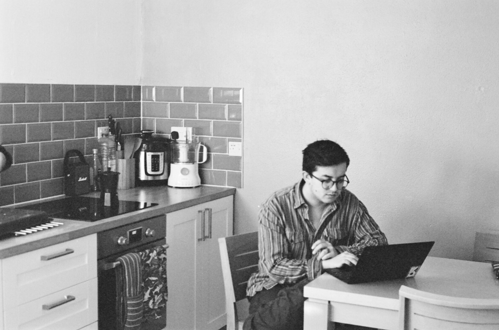 A black and white photograph of a person sat at a laptop at the kitchen table