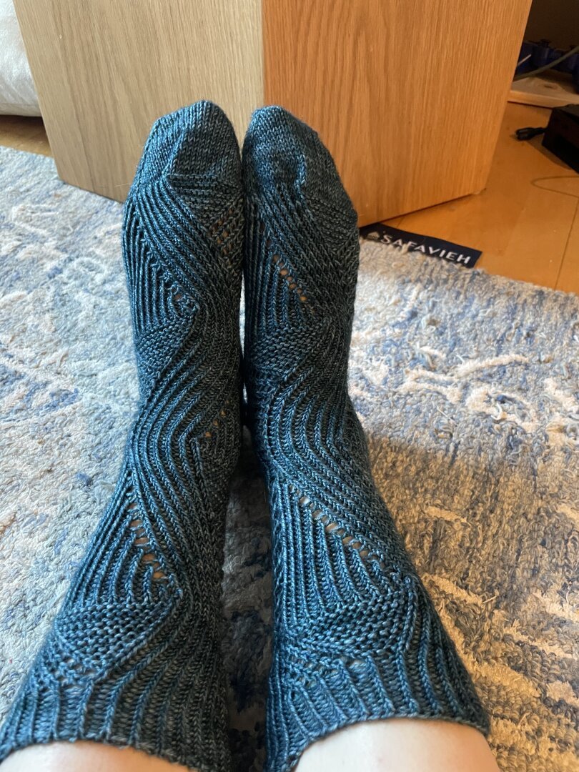 A picture of the top and legs of a pair of knitted socks, on feet. They have vertical waves made by a 1x1 rib and simple lace.